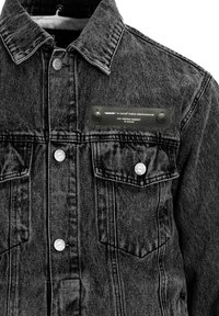 Chaqueta de mezclilla negra con cuello puntiagudo, dos bolsillos en el pecho, herrajes plateados y una etiqueta negra con texto y dos botones a presión.
