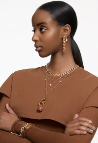 Guld smykker med lagdelte kæder, krystal detaljer og en struktureret vedhæng med røde accenter; båret med en brun top.