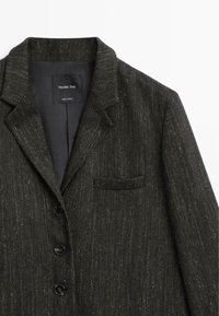 Blazer nero con texture, dotato di rever a punta. Presenta una tasca sul petto, quattro bottoni e un rivestimento interno liscio. Etichetta del brand visibile.
