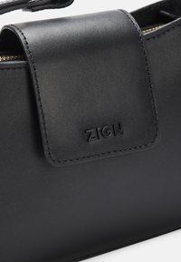 Zwarte leren handtas met een gestructureerd ontwerp, een gladde textuur en een opvallende magneetsluiting met de ingedrukte "ZIGN" logo.