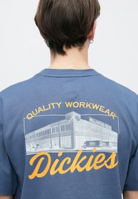 Άτομο με κοντά καστανά μαλλιά που φοράει μπλε πουκάμισο με κίτρινο λογότυπο «Quality Workwear Dickies» και απεικόνιση κτηρίου στην πλάτη.