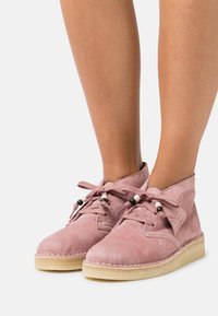 Botas de tobillo de ante rosa empolvado con punteras redondas, cordones de tela y cuentas decorativas. Suela de goma texturizada y detalles cosidos.