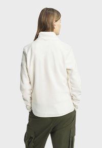 Pull en polaire blanche avec un col montant, des manches longues et un ourlet rond. Détails de couture visibles au niveau du col et des coutures latérales.