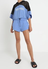 Maglione con cappuccio in rete blu con dettagli neri e logo, abbinato a pantaloni corti azzurri chiari con vita elastica e cordino. Infradito neri.