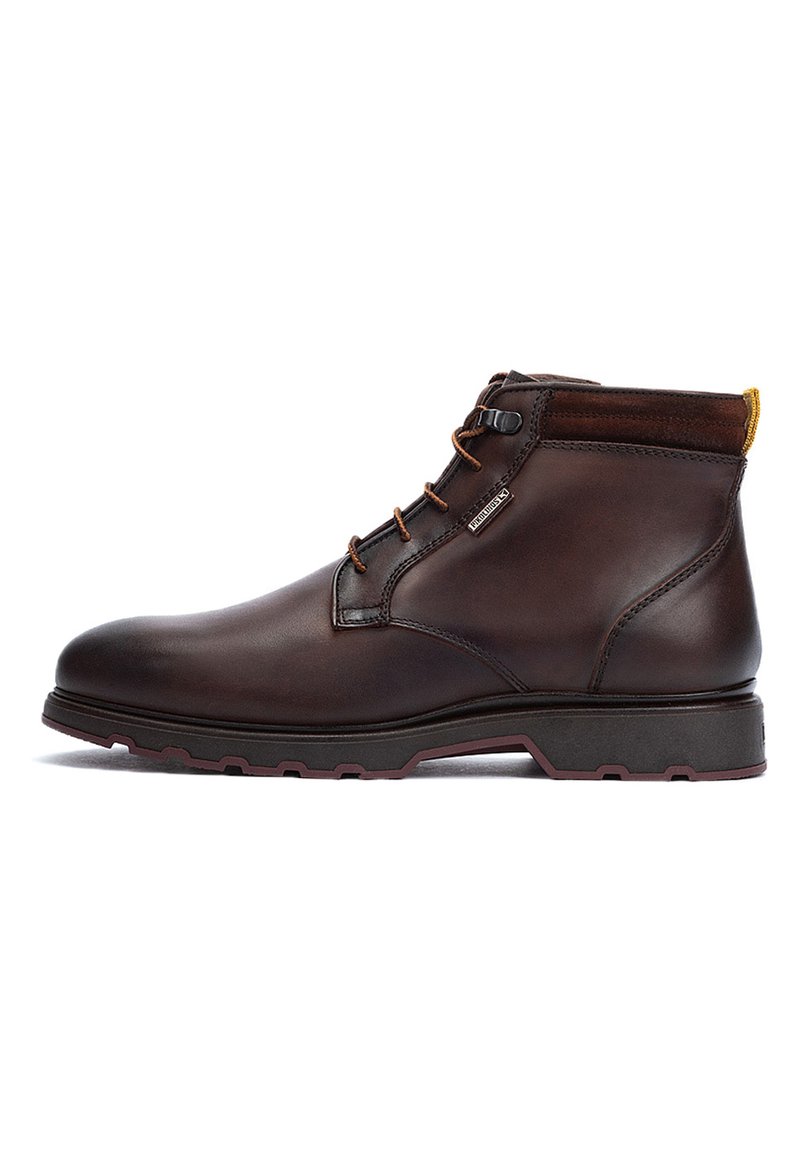 Brown leather ankle boot with a smooth texture, round toe, and rubber sole. Features metal eyelets and a contrasting yellow pull tab.