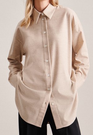 Vrouw draagt een beige overhemd met knopen, lange mouwen en kraag, handen in de zakken, gecombineerd met een zwarte broek tegen een neutrale achtergrond.