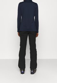 Persona che indossa una giacca blu Navy, pantaloni neri con zip all'orlo e scarponi da sci neri Salomon, in piedi su uno sfondo bianco.
