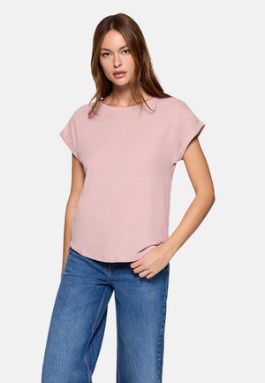 Mujer con cabello largo y castaño, vestida con una camiseta de manga corta color rosa claro con puños enrollados y vaqueros azules, de pie con una mano en el bolsillo.