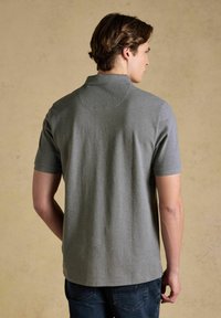 Noor mees, kes vaatab seljataga, kandes lihtsat halli lühikese varrukaga polo särk ja tumedat sinist teksapükse neutraalse beeži taustaga.