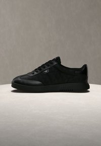 Μαύρο χαμηλό sneaker με δερμάτινες λεπτομέρειες, κορδόνια και ένα διακριτικό λογότυπο στο πλάι, τοποθετημένο σε ανοιχτή επιφάνεια με ουδέτερο φόντο.