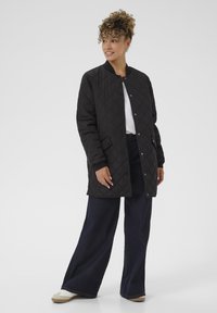 Giubbotto nera trapuntato con chiusura a bottoni e tasche laterali, abbinato a pantaloni in denim scuri a gamba larga e scarpe di colore chiaro.