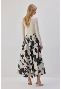 Roomkleurige top met lange mouwen in ribbreisel, gecombineerd met een volle rok tot op de kuit met een zwart-wit abstract patroon. De hakken zijn lichtbeige.