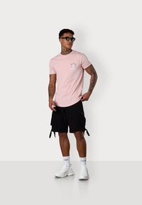 Light pink short-sleeve t-shirt features a front graphic. Black cargo shorts with side pockets. White chunky sneakers and black sunglasses.