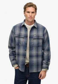 Giovane uomo che indossa una giacca a quadri blu e grigia sopra un maglione beige, con jeans scuri e cintura di pelle marrone, in piedi contro uno sfondo bianco.