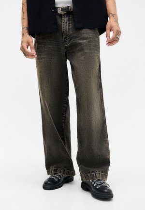 Jean en denim gris foncé à jambes larges avec une texture délavée. Porté avec une chemise blanche et une ceinture noire, agrémentée de ferrures en argent et de tatouages visibles.