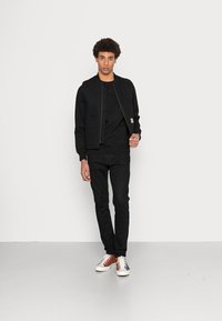 Giacca bomber in cotone nera con chiusura a zip, abbinata a una maglietta nera a girocollo e jeans neri. Le sneakers presentano dettagli rossi e blu.