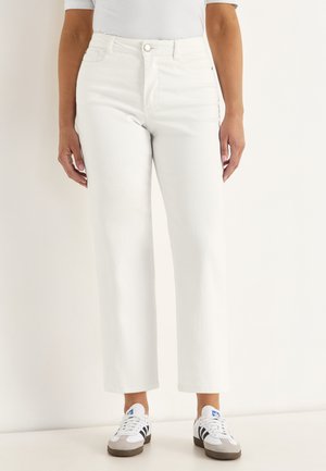 Person iført hvide bukser med høj talje og lige ben samt hvide sneakers med sorte striber, stående mod en ensfarvet lys baggrund.