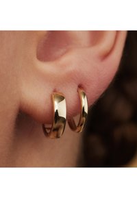 Goldene Creolen, die an einem durchstochenen Ohr befestigt sind, mit einer glatten, glänzenden Oberfläche und einem polierten Design. Details der Krümmung sind sichtbar.