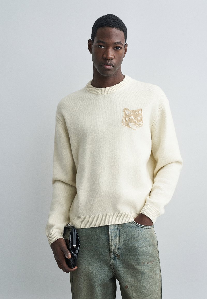 Férfi krémszínű pulóvert visel, amelyen egy bézs róka arc részlet látható, megfakult zöld farmernadrágot hord, és egy fekete texturált táskát tart egy semleges stúdióbeállításban.