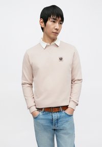 Lyserosa genser med rund hals og en hvit skjorte med krage under. Har en liten logopatch på brystet. Brukes sammen med blå jeans.