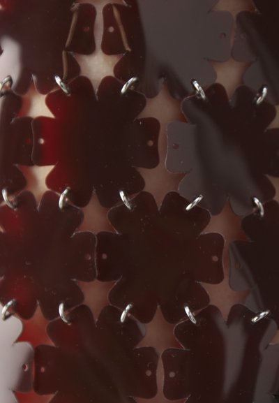 Red flower-shaped elements made of glossy material, linked with small metal rings, creating a layered pattern with visible cut-outs.
