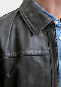 Chaqueta de cuero negra con una textura suave, que presenta un cuello clásico, cierre de cremallera y un brillo sutil. Superpuesta sobre una camisa azul.