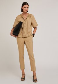 Blusa beige a maniche corte con scollo arricciato e chiusura con bottoni, abbinata a pantaloni beige aderenti e borsa nera a spalla. Accentata da un orologio.