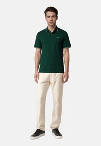 Zielona koszulka polo z białymi akcentami, krótkim rękawem; zestawiona z beżowymi spodniami i ciemnymi sneakersami. Gładki materiał, klasyczny krój kołnierzyka.