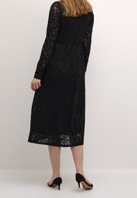 Svart spetsklänning med långa ärmar och figurnära livstycke, som flyter ner till en midkalvslängd. I kombination med svarta högklackade skor.