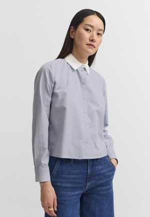 Woman with long dark hair wearing a light blue buttoned shirt with white collar and blue jeans standing against a plain background.