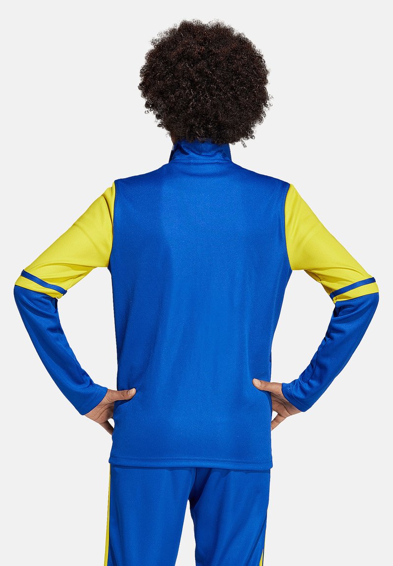 Personne aux cheveux bouclés, debout les mains sur les hanches, portant une tenue de sport manches longues bleue et jaune, vue de dos sur un fond blanc.