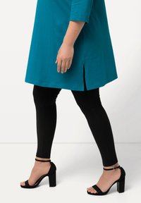Teal tunika med trekvartsärmar och sidoklyftor, kombinerad med svarta leggings och svarta ankelremmar. Slätt tyg, avslappnad passform.
