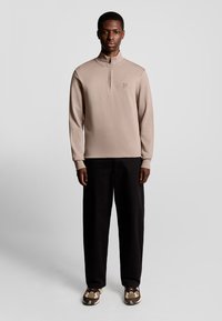 Beige half-zip trui gemaakt van zachte stof, gecombineerd met zwarte wijd-uitlopende broeken en gestepte schoenen met bruine en crème accenten.