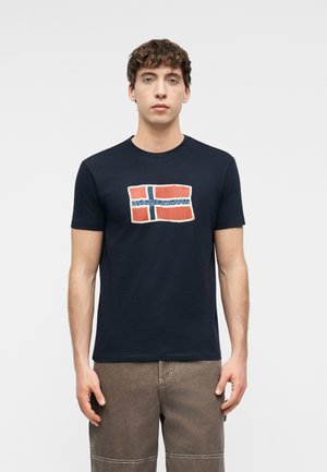 Fiatal férfi, sötétkék pólóban, amelyen piros, fehér és kék stilizált zászlóminta látható, barna nadrágban, egyszínű háttér előtt áll.