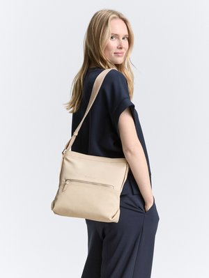 Femme blonde en tenue bleu marine portant un sac bandoulière beige avec une poche zippée à l'avant, regardant par-dessus son épaule sur un fond uni.