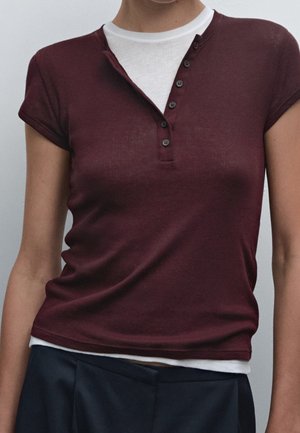 Femme portant un t-shirt henley bordeaux à manches courtes superposé à un tee-shirt blanc avec un pantalon bleu marine, montrée des épaules aux hanches sur un fond uni.