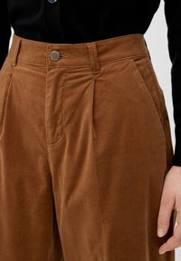 Hnědé kordové kalhoty s předními sklady, zapínáním na knoflík a bočními kapsami, nošeny s černým topem; ruka spočívá u boků.