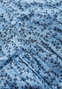 Blauer Stoff mit einem Blattmuster in Dunkelblau und Weiß, mit einem kleinen blauen Knopf und einer Faltendetails in der Mitte.