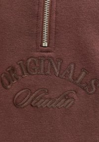 Sweat à zip couleur bordeaux avec un logo brodé "ORIGINALS" et "Studio" en fil marron, texture de tissu lisse, fermeture éclair métallique.