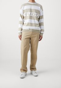 Pull rayé beige et blanc avec le logo "HUGO" au centre, associé à un pantalon beige et des baskets blanches, se tenant devant un fond neutre.