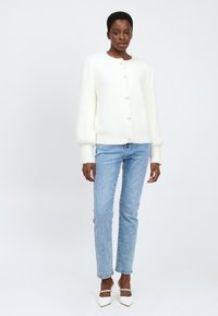 Vrouw in een witte, gebuttonde, fuzzy trui, lichtblauwe straight-leg jeans en witte hakken met enkelbandjes tegen een effen achtergrond.