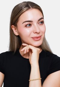 Zlatna narukvica s kružnim akcentima, nosi se na zapešću. Model ima dugu kosu, neutralan make-up i pozira s promišljenim izrazom lica.