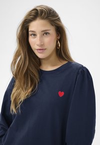 Marineblaues Langarmshirt aus weichem Stoff mit rundem Ausschnitt und Puffärmeln, versehen mit einer kleinen roten Herzstickerei auf der Brust.