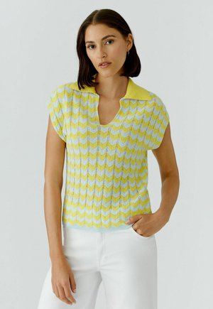 Femme aux cheveux foncés mi-longs portant un haut sans manches en maille à motif chevron jaune et blanc et un pantalon blanc, debout devant un fond uni.
