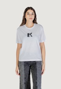 T-shirt bianco in cotone con maniche corte, caratterizzato da un design del logo stampato in nero sulla parte anteriore. Abbinato a jeans a gamba larga grigio scuro.