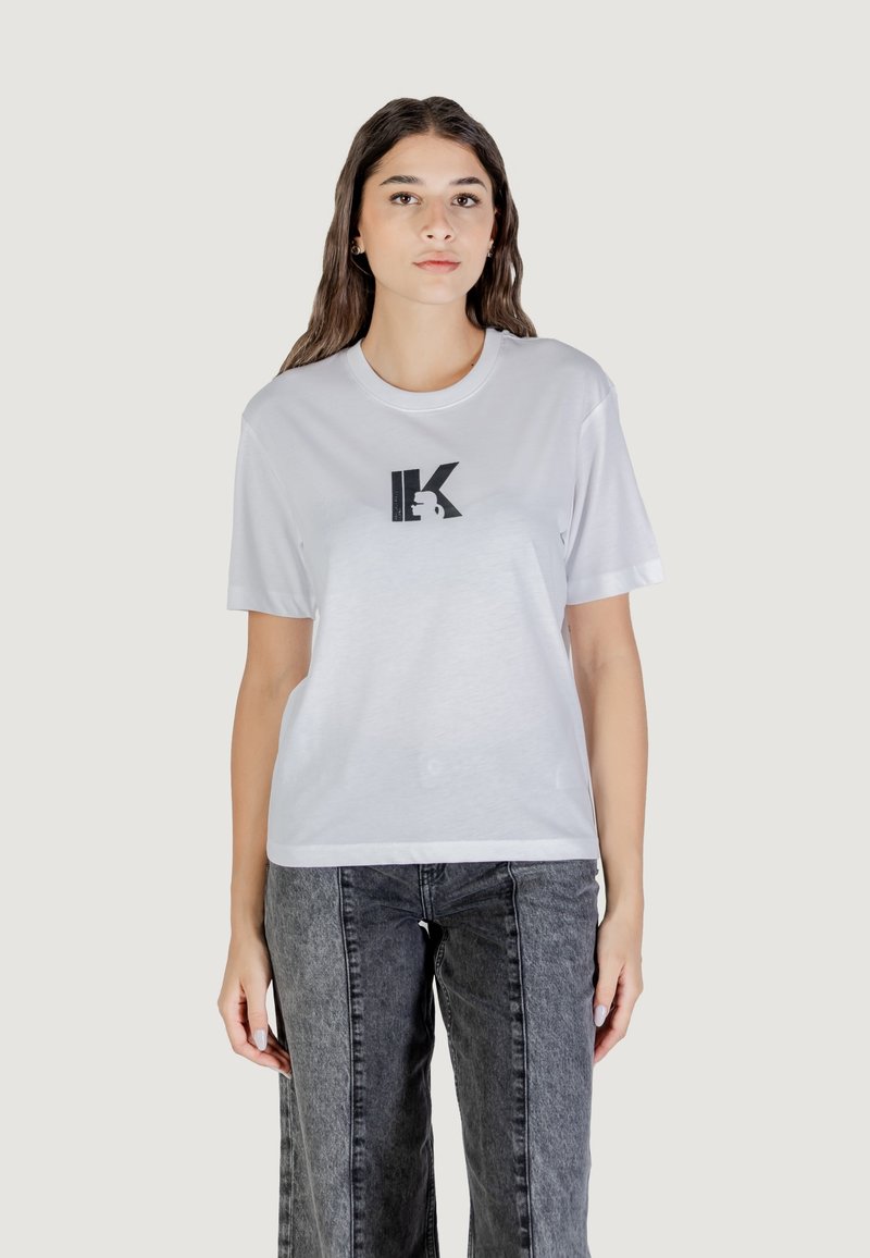 T-shirt bianco in cotone con maniche corte, caratterizzato da un design del logo stampato in nero sulla parte anteriore. Abbinato a jeans a gamba larga grigio scuro.