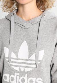 Šedá mikina s kapucí vyrobená z měkké tkaniny, s velkým bílým logem Adidas a pruhovaným vzorem. Obsahuje odpovídající šňůrku a kovové detaily.