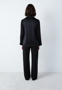 Camisa negra de satén de manga larga con cuello, combinada con pantalones sueltos a juego; textura suave, diseño minimalista y sin patrones visibles.