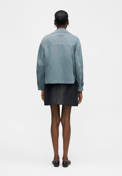 Femme à la peau foncée et aux cheveux attachés en chignon, portant une veste en jean bleu clair, une jupe noire, des boucles d'oreilles créoles et des ballerines noires, vue de dos.