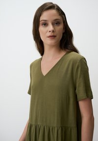Olive green dress made of textured fabric, featuring a V-neck, short sleeves, and a flared, tiered bottom. Simple and loose-fitting design.